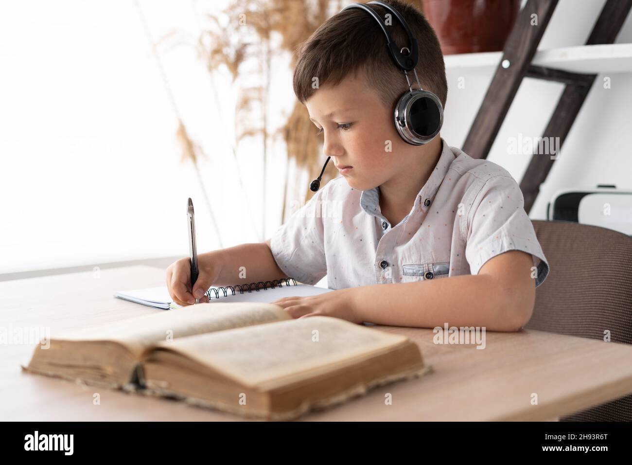 L'allievo caucasico in cuffie alla scrivania facendo i compiti in un notebook legge un libro di testo. Il bambino ha una lezione di auto-isolamento a casa. Online Foto Stock