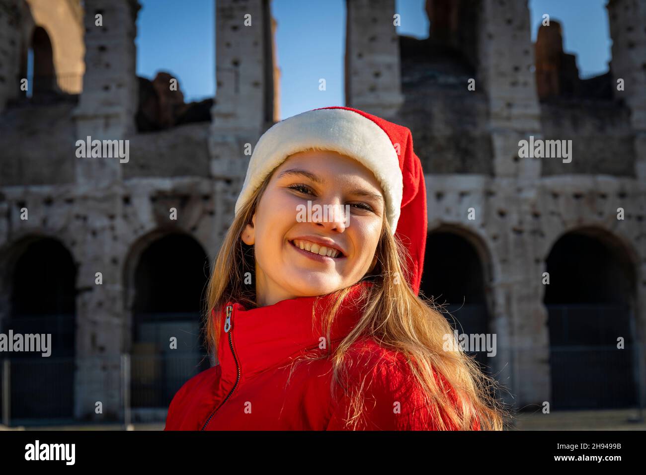 Vacanze romane a Natale. Bella donna bionda in posa davanti al Colosseo con cappello di Babbo Natale. La giovane donna guarda nella macchina fotografica e s Foto Stock