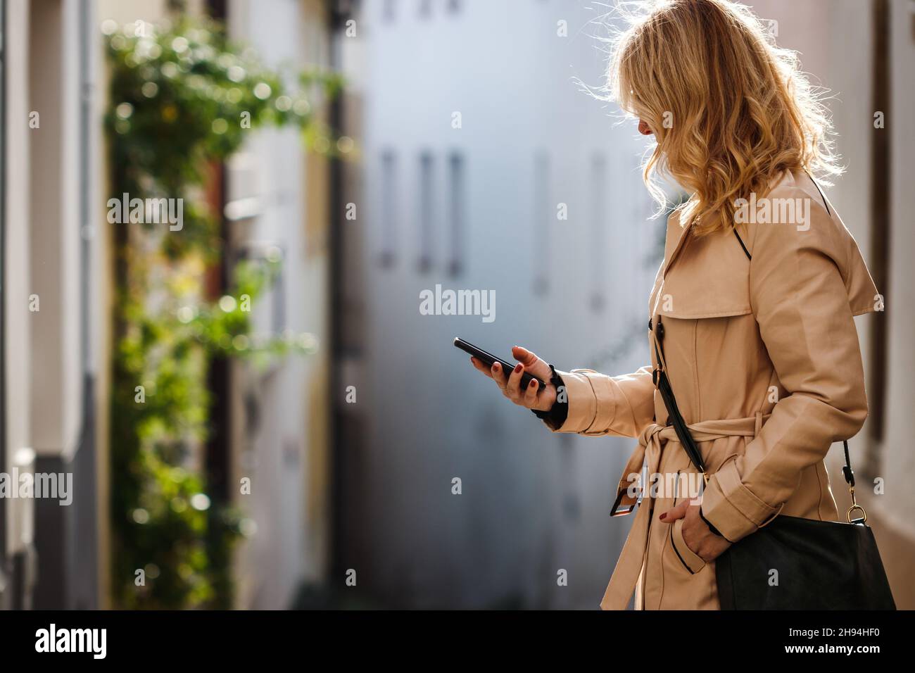 Donna con trench che usa il suo smartphone sulla strada della città. Messaggi di testo per donna sul telefono cellulare Foto Stock