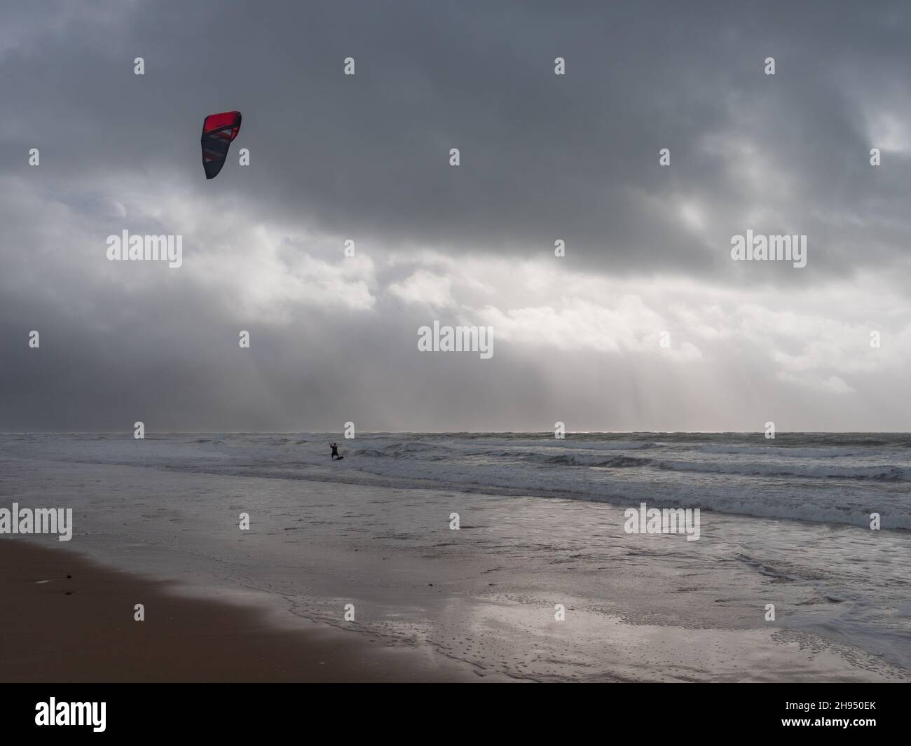 Un kitesurfer surf lungo le onde su una spiaggia di sabbia contro un cielo nuvoloso e tempestoso in Galles Foto Stock