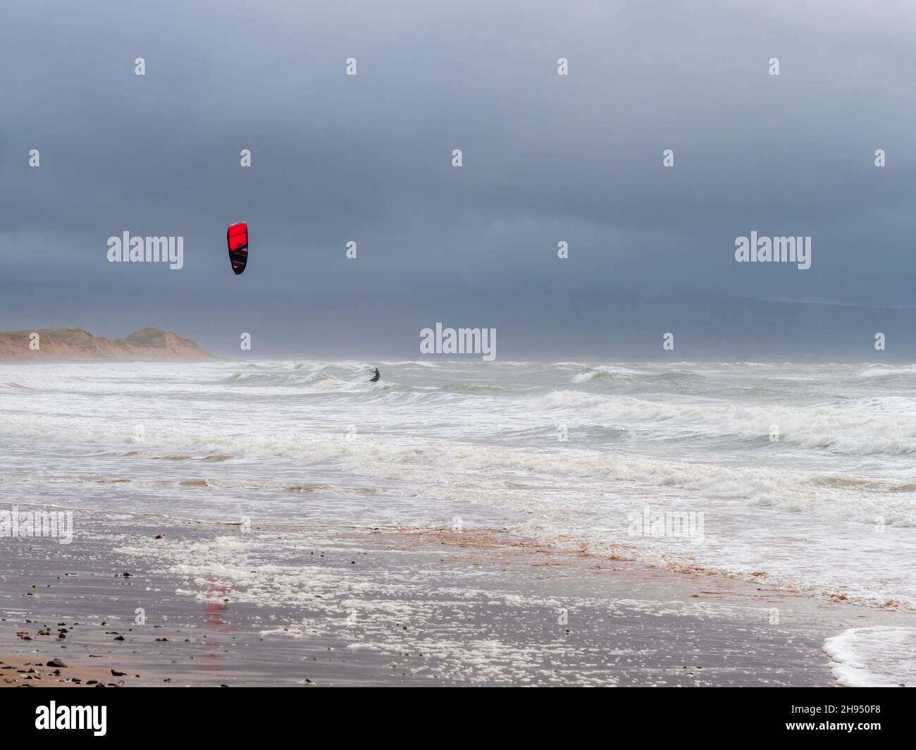 Un kitesurfer surf lungo le onde su una spiaggia di sabbia contro un cielo nuvoloso e tempestoso in Galles Foto Stock