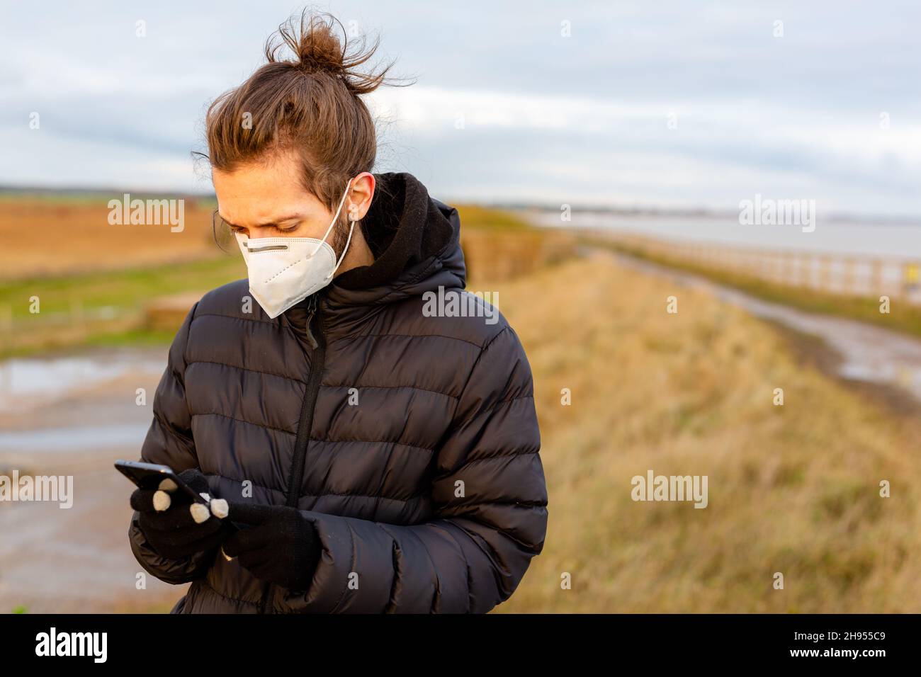 Un giovane uomo che indossa una maschera facciale KN95 One use mentre usa il suo cellulare durante la pandemia del virus della corona Covid-19 Foto Stock