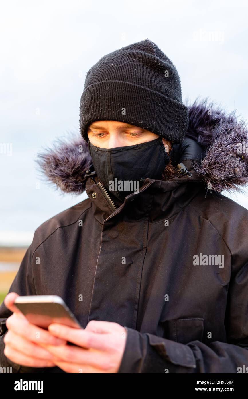 Un giovane uomo che indossa una maschera nera lavabile mentre usa il suo cellulare durante la pandemia del virus della corona Covid-19 Foto Stock