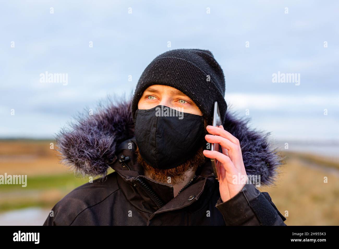 Un giovane uomo che indossa una maschera nera lavabile mentre usa il suo cellulare durante la pandemia del virus della corona Covid-19 Foto Stock