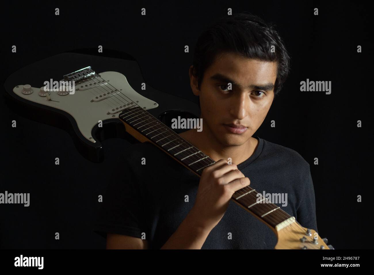 giovane e attraente musicista rock latino che posa con la sua chitarra elettrica sopra la spalla all'interno di uno studio con sfondo nero Foto Stock