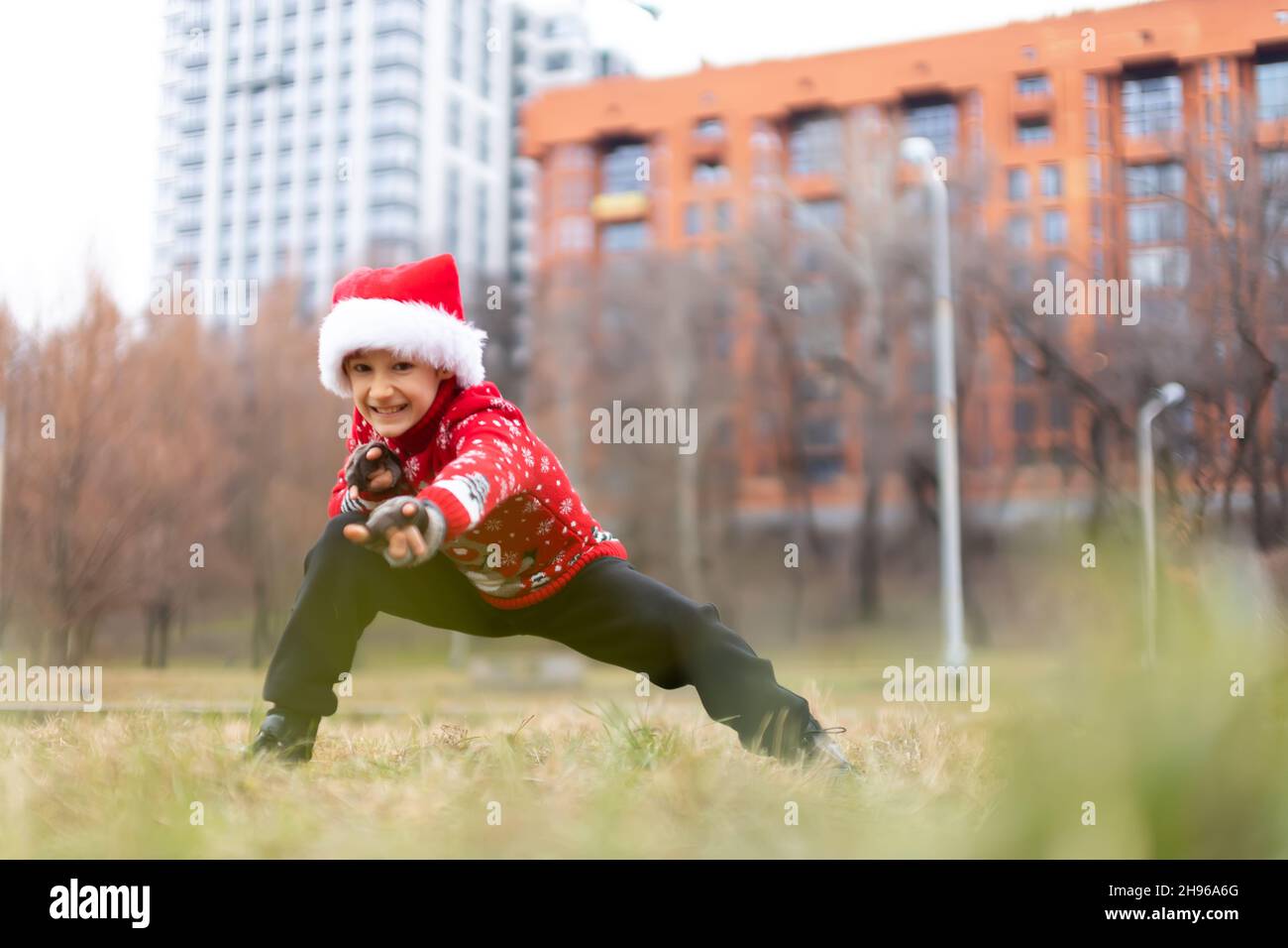 Un ragazzo in un caldo maglione di Natale con un cervo di Capodanno e un cappello Santa esegue esercizi di stretching e ginnastica mentre si fanno i volti Foto Stock