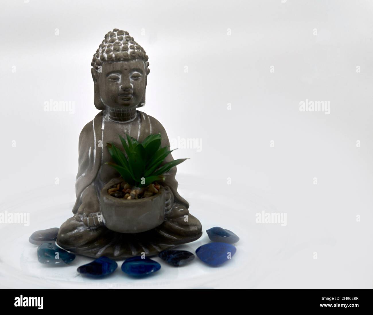 Un piccolo Buddha grigio con un succulento circondato da sette pietre blu semi-preziose in acqua. Sfondo bianco. Foto Stock