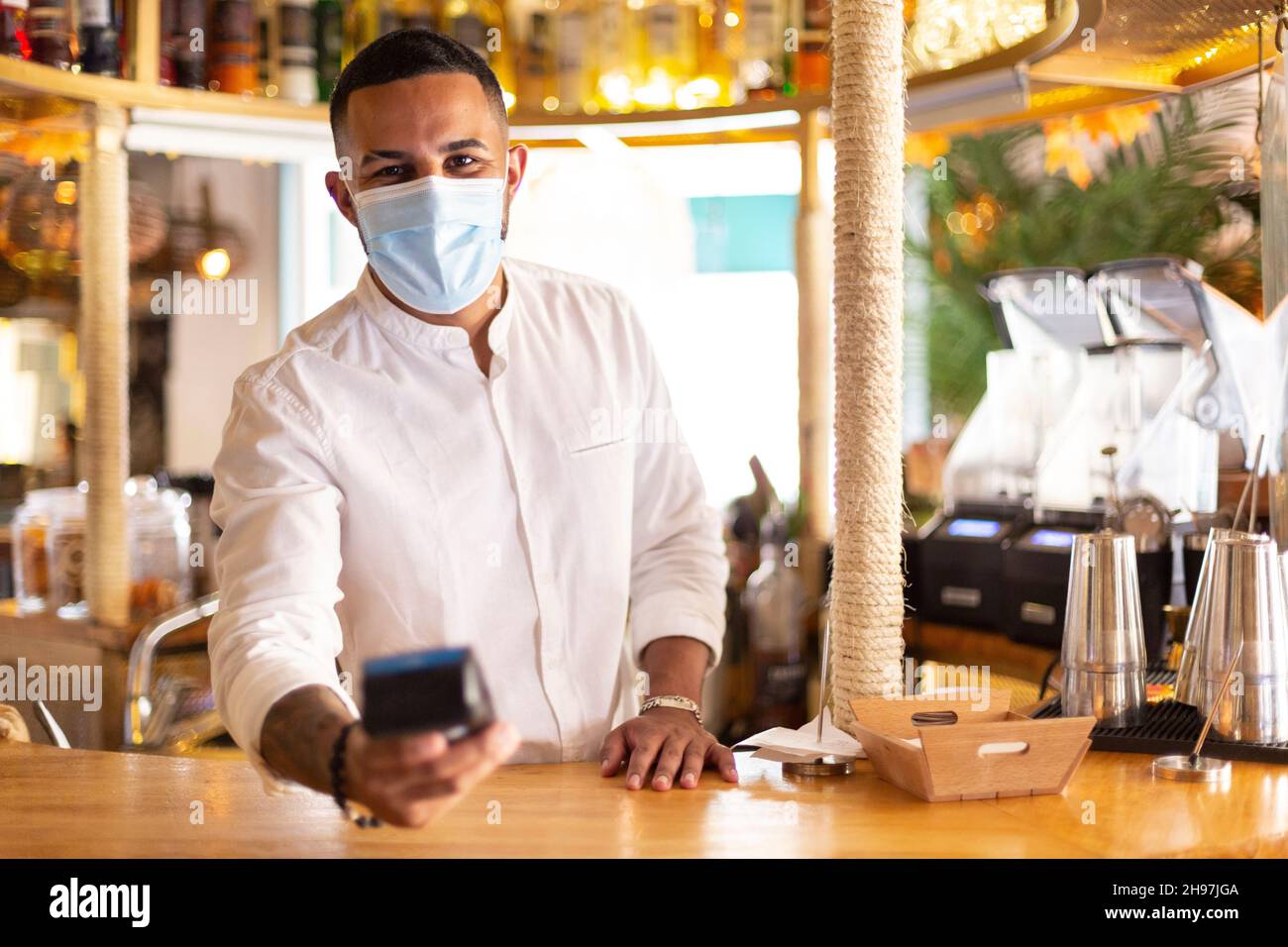 Elegante cameriere latino-americano con un dataphone in mano per effettuare un pagamento. Indossa una maschera medica. Spazio per il testo. Foto Stock