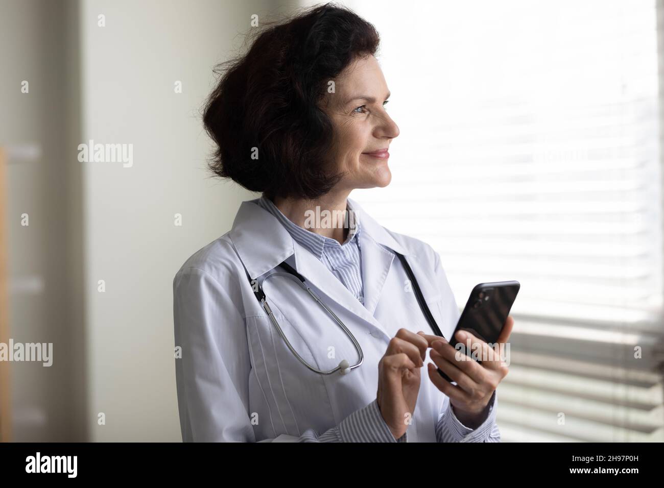 Felice pensiva donna medico di mezza età utilizzando l'app online Foto Stock