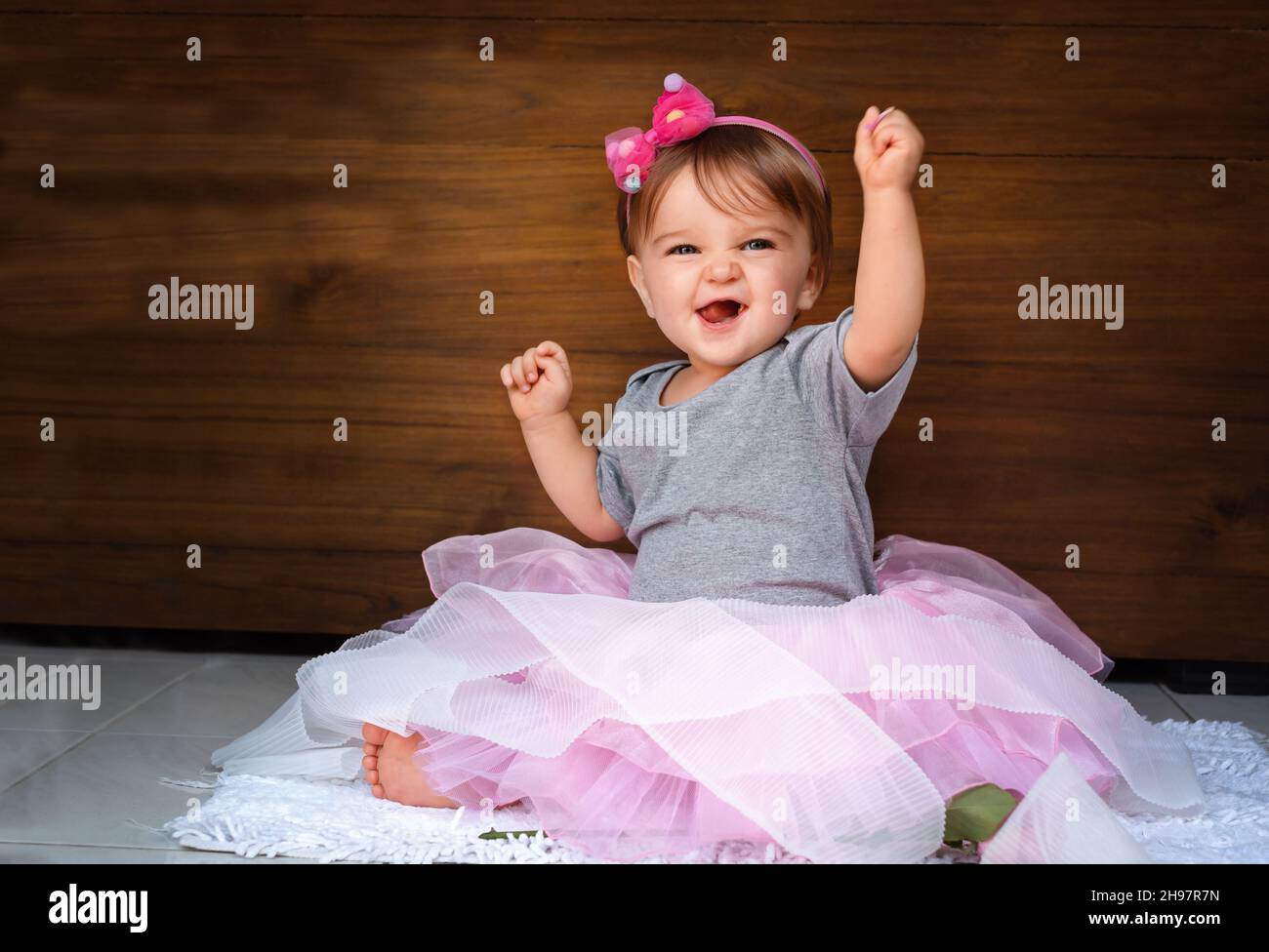 Il bambino urla con gioia. Ragazza bambino felice nella gonna rosa sullo sfondo di legno sollevato le mani e ride felicemente Foto Stock
