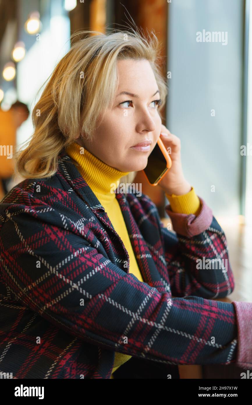 Una bella bionda caucasica di mezza età sta parlando sul telefono cellulare in un bar. Foto Stock