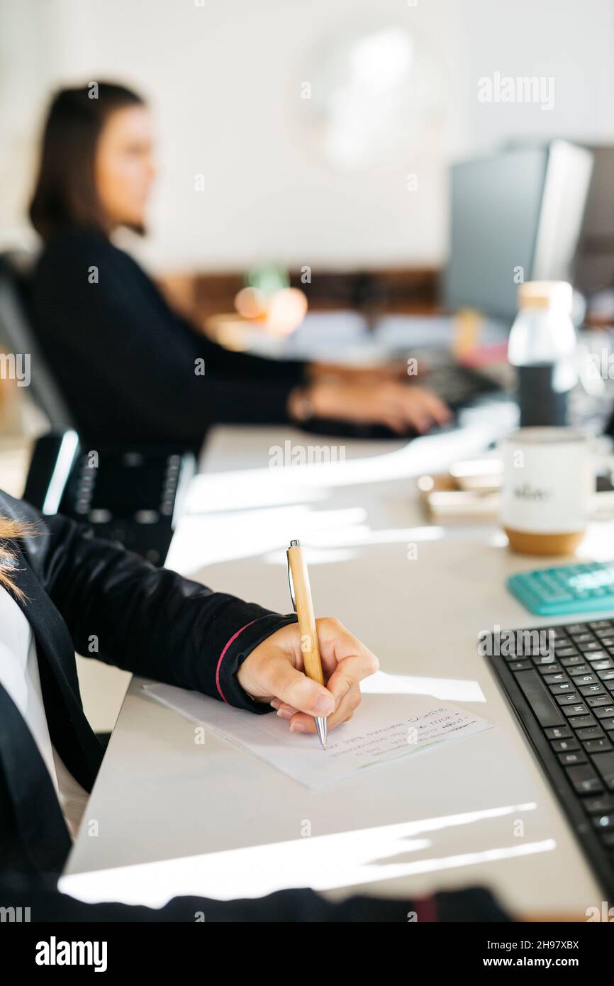 Primo piano delle mani della donna che scrivono con una penna su una scrivania disordinata in un ufficio Foto Stock