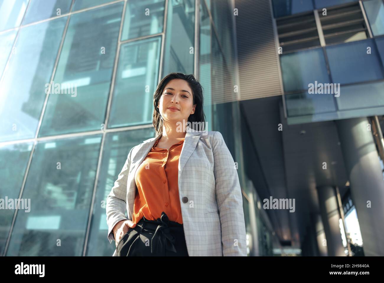 Donna d'affari sicura che guarda la macchina fotografica mentre si trova di fronte a un alto edificio di uffici in città. Imprenditore femminile di successo stand Foto Stock
