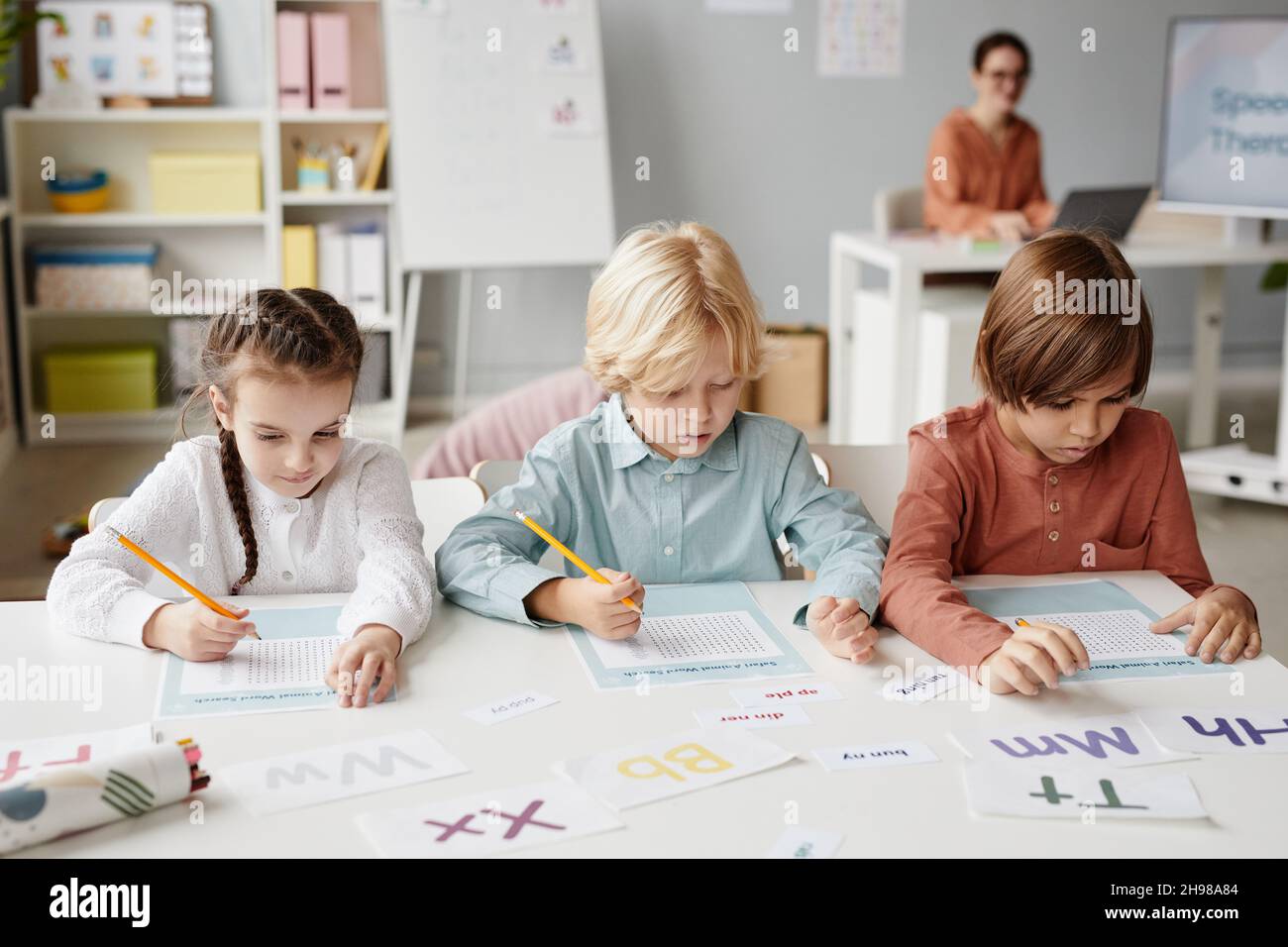 Gruppo di bambini della scuola seduti al tavolo e prendere appunti sui documenti durante la lezione a scuola con l'insegnante in background Foto Stock