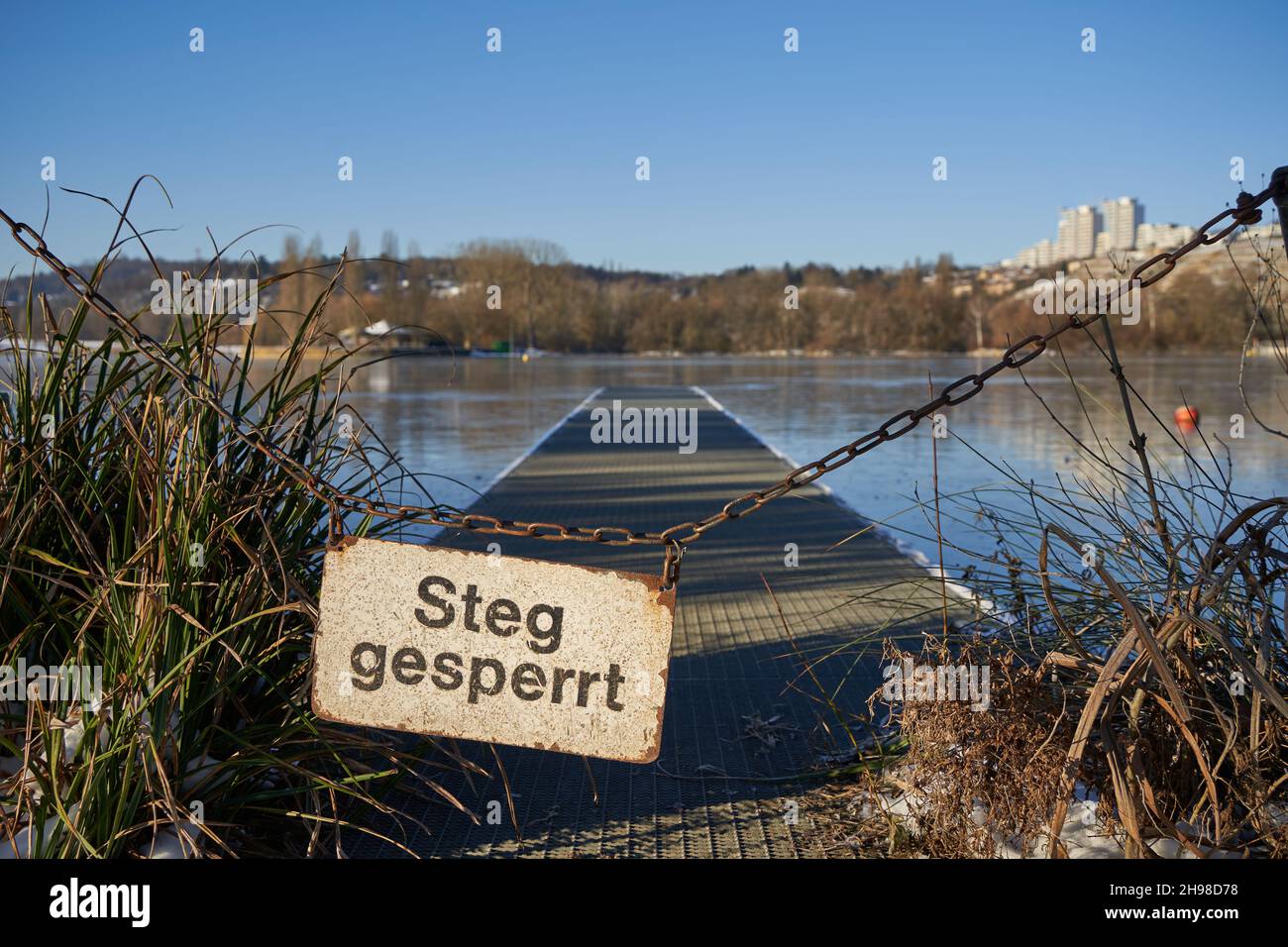 Firma su una catena arrugginita dicendo: Dock bloccato ( tedesco: Steg gesperrt). Lettere nere su lavagna bianca. Lago blu congelato e grattacieli sullo sfondo, Foto Stock