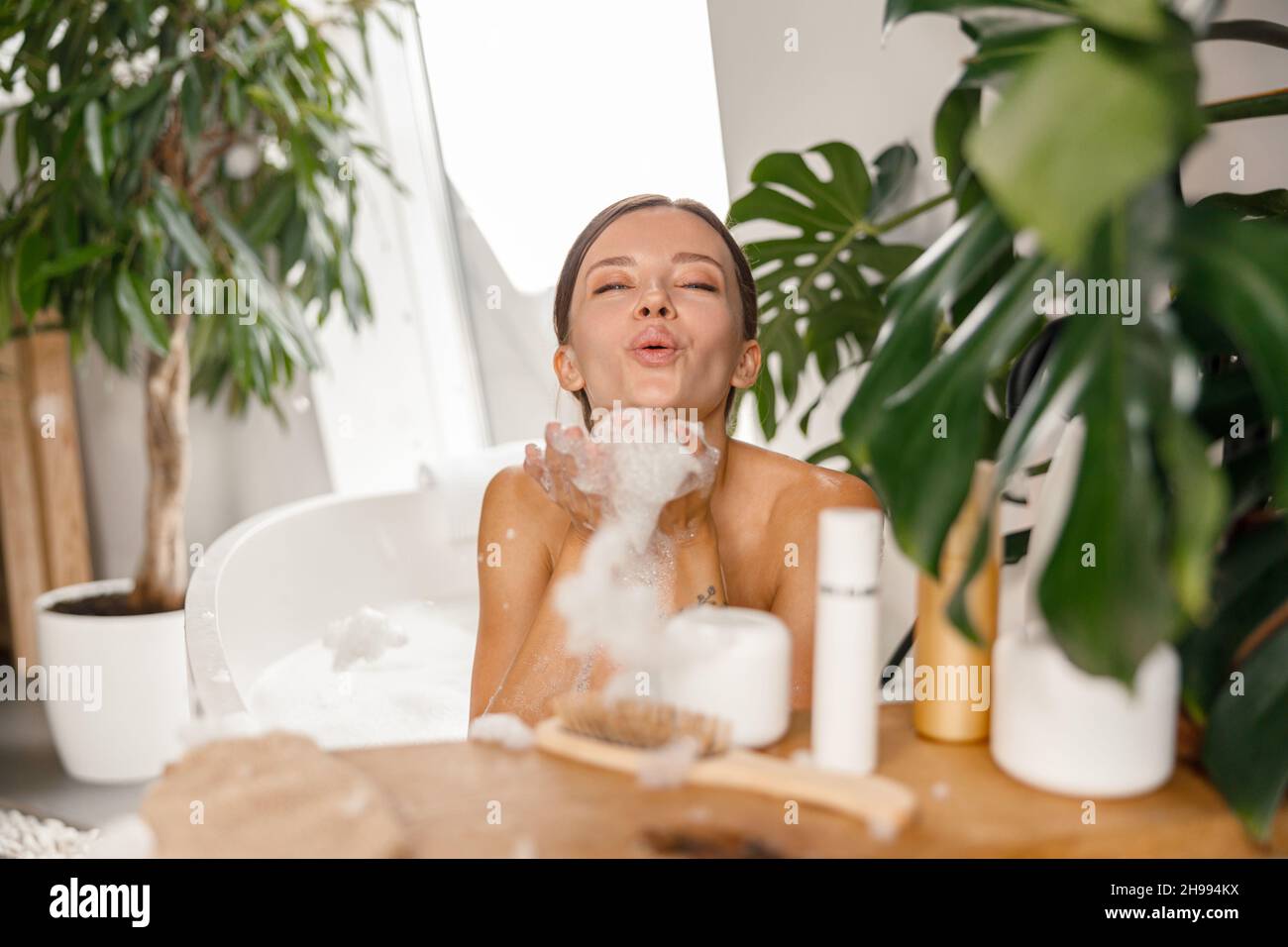 Una giovane donna giocosa che soffia schiuma di sapone mentre fa il bagno a bolle presso il centro benessere Foto Stock