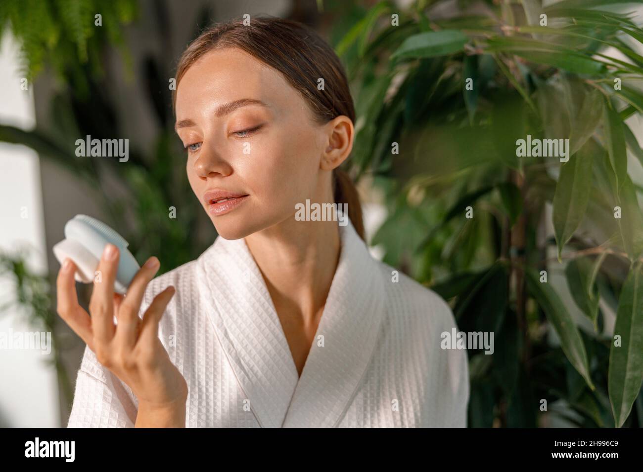 Giovane donna in accappatoio cercando di utilizzare la spazzola viso in silicone per esfoliazione della pelle, avendo routine di bellezza in bagno decorato con piante Foto Stock