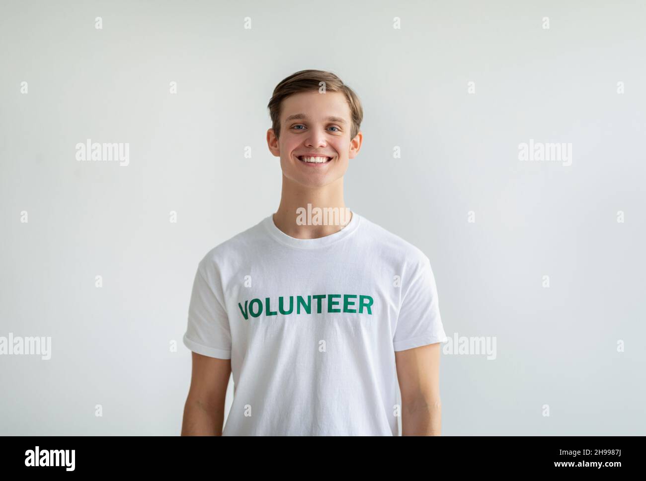 Felice giovane ragazzo volontario in t-shirt bianca in piedi su sfondo chiaro e sorridente a macchina fotografica, ritratto studio Foto Stock