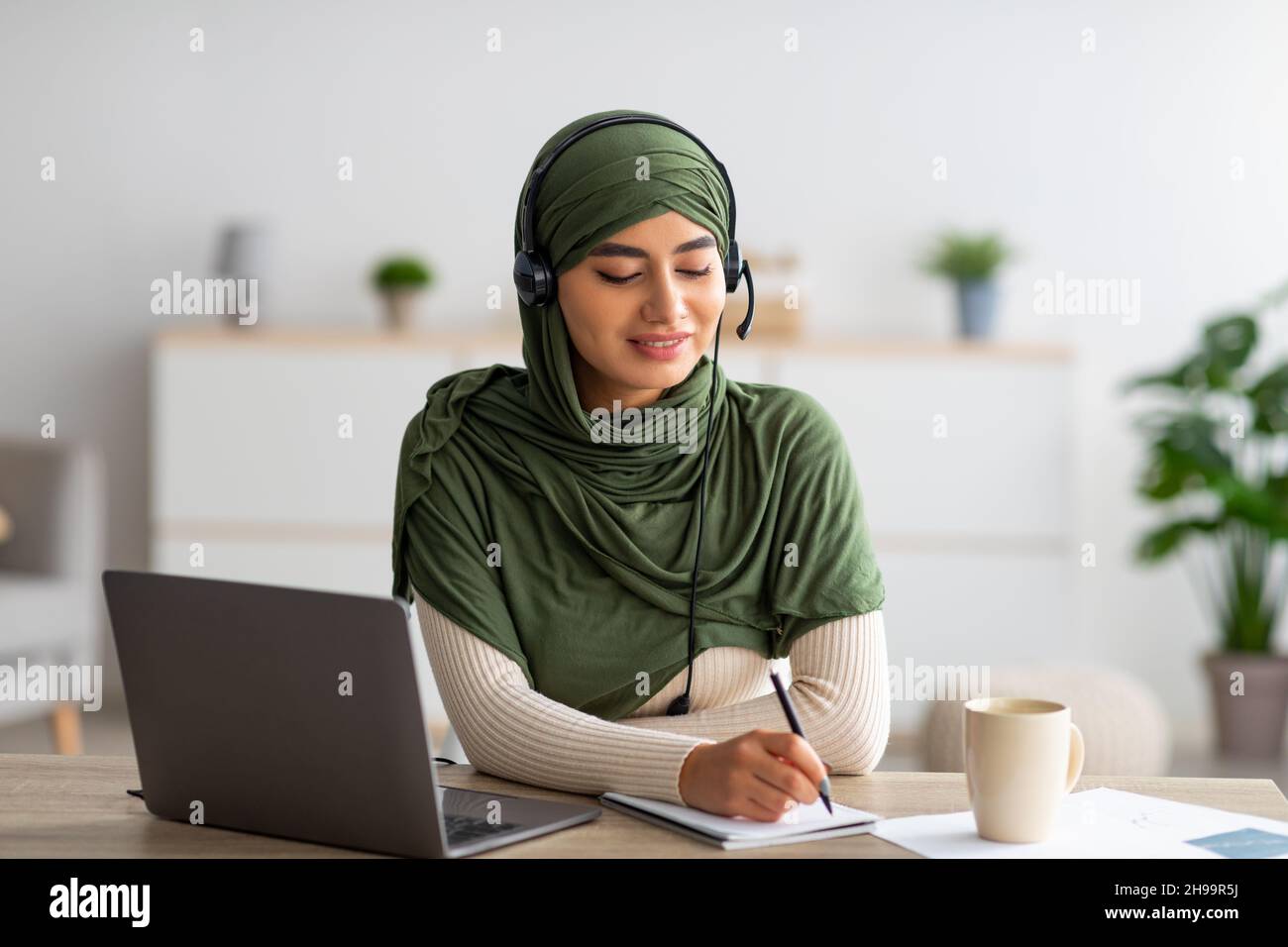 Giovane donna araba in hijab che lavora su un notebook, indossa le cuffie, prende appunti durante il webinar a casa Foto Stock