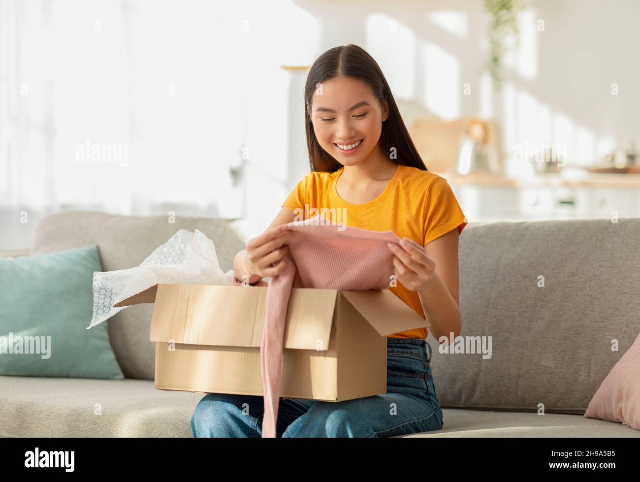 Eccitata giovane donna asiatica che apre la scatola di cartone, guardando nuovi vestiti e sorridendo, soddisfatto della sua consegna Foto Stock