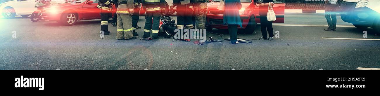 Un sacco di vigili del fuoco, paramedici e soccorritori al posto di incidente in bicicletta e incidente d'auto, rotto e relitto moto su strada. Ampia immagine panoramica. Foto Stock