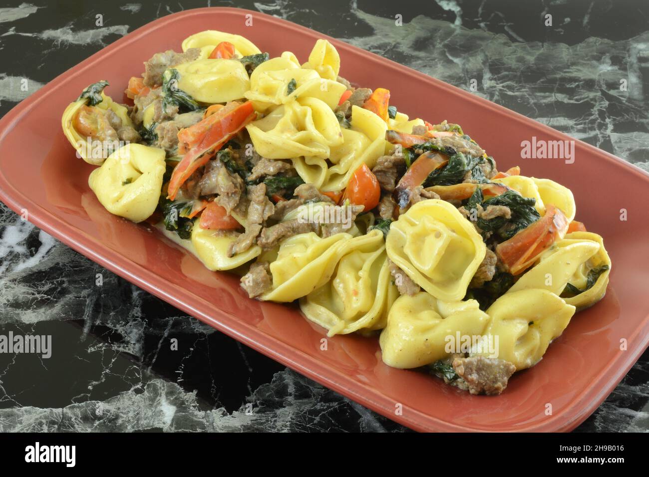 Tortellini al parmigiano con spinaci, bistecche e pomodori cena su piatto rosso Foto Stock