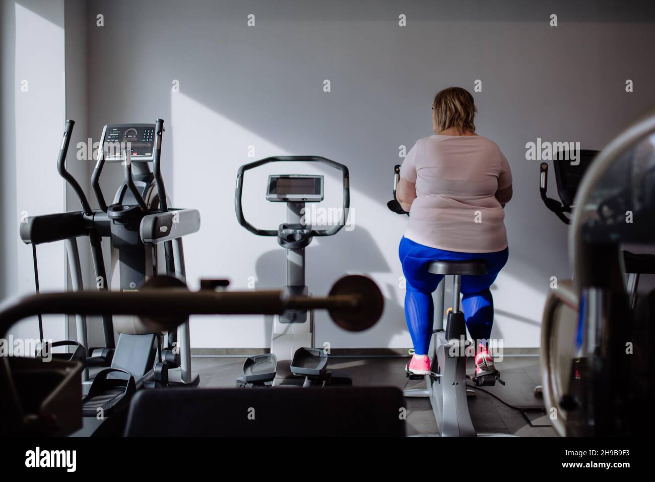 Vista posteriore della donna di peso medio adulto che si esercita sulla cyclette interna in palestra Foto Stock