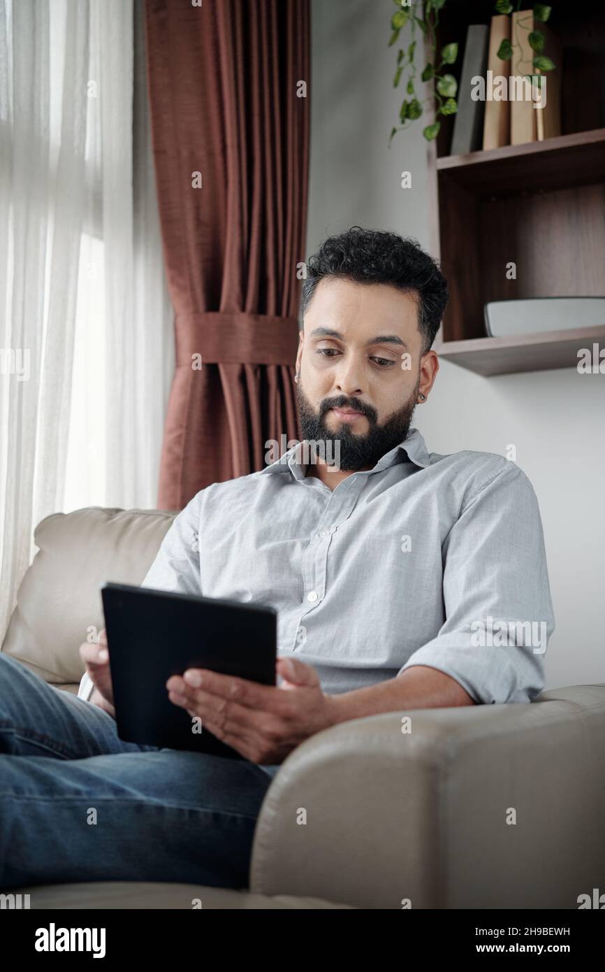 Positivo bel barbuto uomo seduto in poltrona a casa e lettura libro o articolo su tablet computer Foto Stock