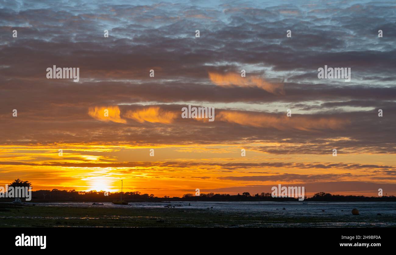 Cielo arancione nuvoloso mentre il sole tramonta su Bosham Quay nel porto da Bosham Village a bassa marea in inverno nel Sussex occidentale, Inghilterra, Regno Unito. Foto Stock