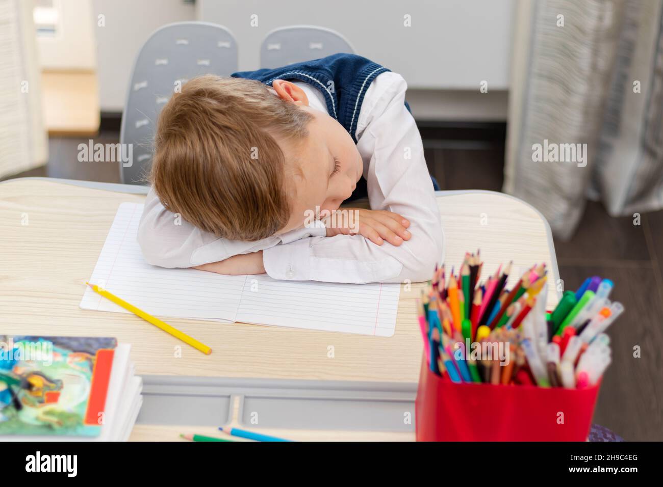 Un ragazzo carino primo grader in una divisa della scuola a casa durante una pandemia si addormentò facendo i compiti ad una scrivania con libri e matite. Messa a fuoco selettiva. Cl Foto Stock