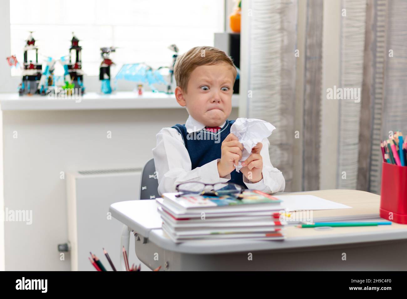 Carino ragazzo prima livellatore in uniforme scuola a casa durante una pausa folgorante mentre si siede alla sua scrivania. Messa a fuoco selettiva. Primo piano. Verticale Foto Stock