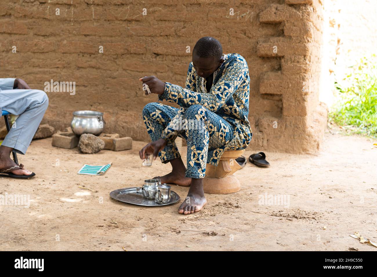 Giovane villager africano che prepara il tè seduto appeso su un piccolo sgabello nel suo cortile Foto Stock