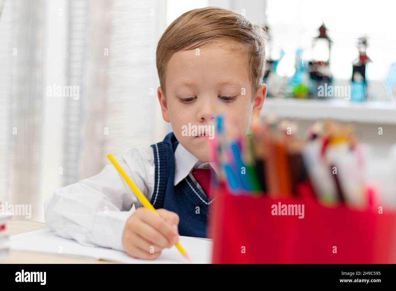 Un ragazzo carino prima livellatrice in una divisa della scuola fa i compiti mentre si siede ad una scrivania con una matita nelle sue mani a casa. Istruzione remota. Focu selettivo Foto Stock