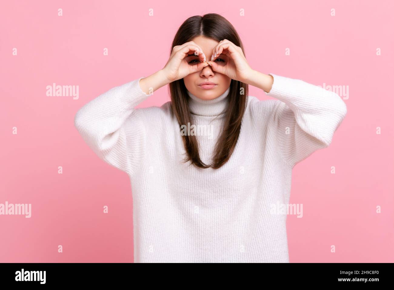 Ritratto di attraente bruna femminile in piedi braccia sollevate e guardare con binocolo gesto, indossando bianco casual stile maglione. Studio interno girato isolato su sfondo rosa. Foto Stock