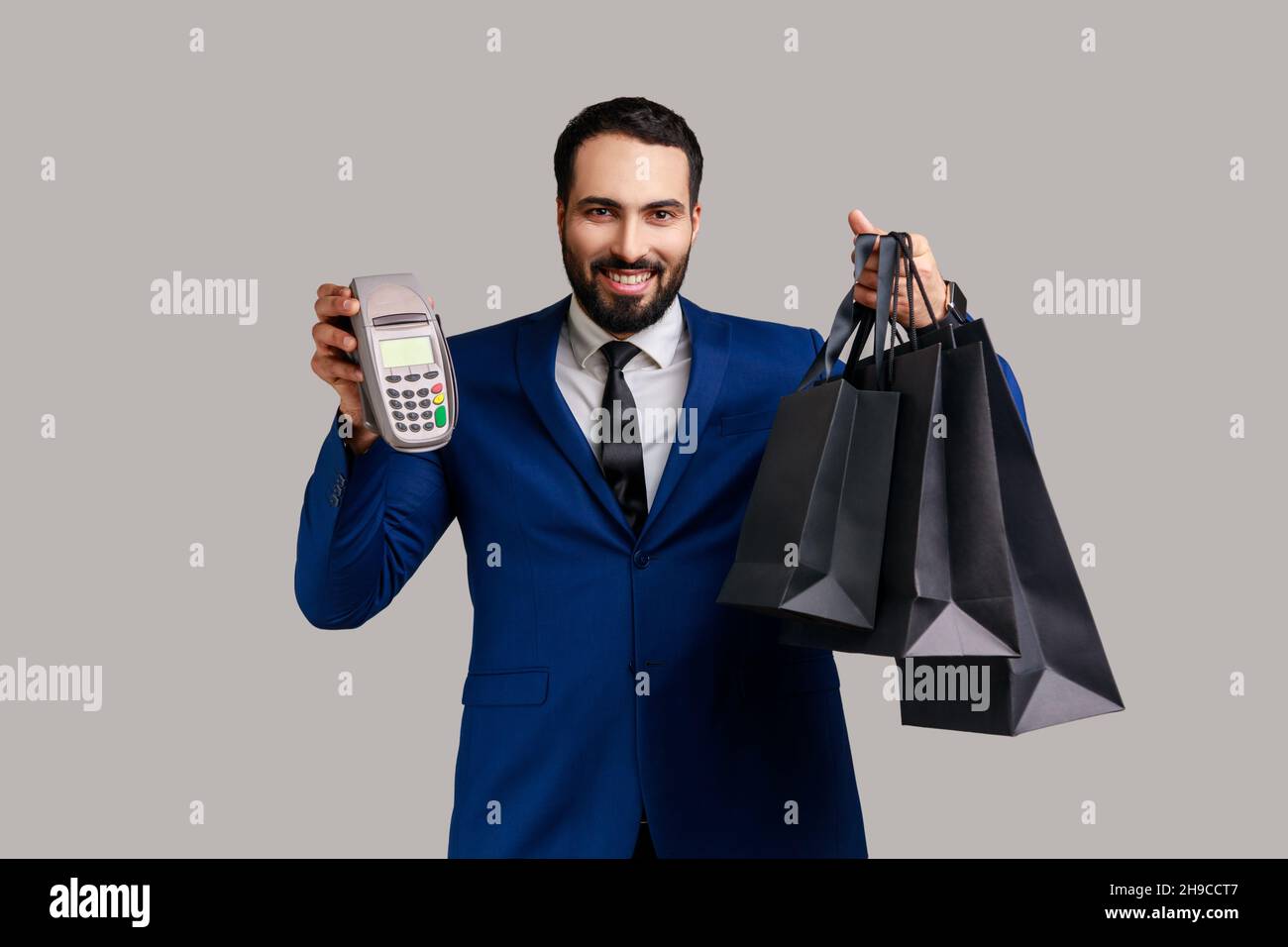 Un uomo d'affari soddisfatto che tiene il terminale di pagamento e le borse di shopping di carta, ordine e consegna espressi facili, vestito di stile ufficiale. Studio interno girato isolato su sfondo grigio. Foto Stock