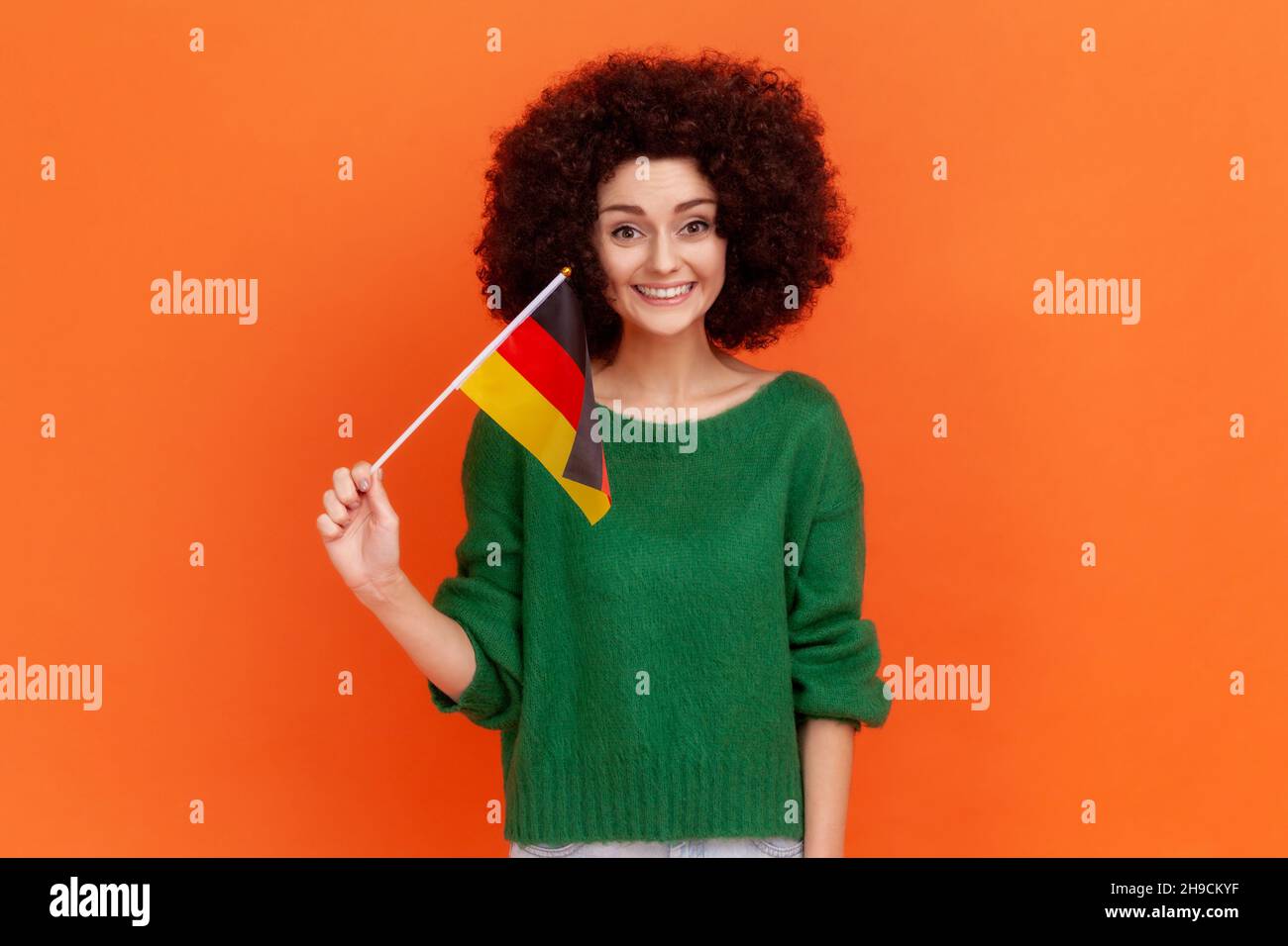 Ritratto di attraente donna positiva con acconciatura Afro indossando verde casual stile maglione con bandiera tedesca, celebrando il giorno dell'unità tedesca. Studio interno girato isolato su sfondo arancione. Foto Stock