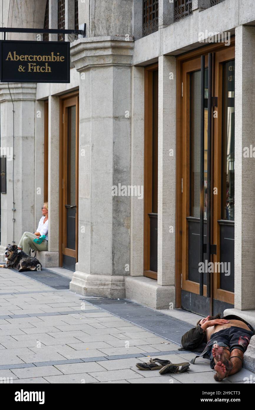 Un uomo senza tetto dorme sul pavimento di fronte al negozio Abercrombie & Fitch a Monaco. Al lato siede una signora con il suo cane. Foto Stock