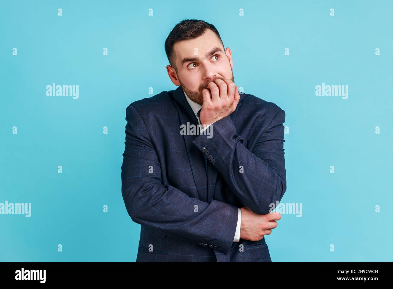 Un uomo d'affari ansioso e nervoso con barba in vestito di stile ufficiale che morde le unghie sulle dita guardando la macchina fotografica con espressione terrorizzata, preoccupato per il lavoro. Studio interno girato isolato su sfondo blu. Foto Stock