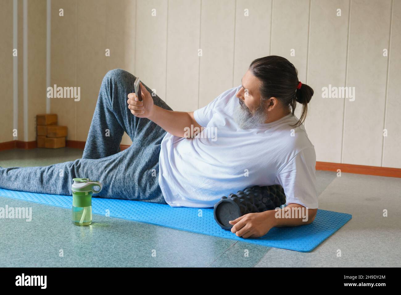 Caucasico bearded, uomo dai capelli lunghi con rullo di schiuma e shaker con acqua che posano sul tappeto yoga e usando il suo telefono cellulare. Concetto di formazione online. Foto Stock