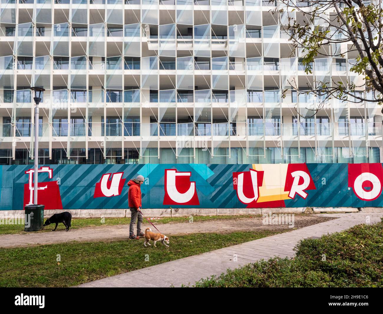 Milano, Lombardia, Italia. Dicembre 2019. Un giovane uomo in giacca rossa cammina il suo cane di fronte ad un edificio con un grande segno con la frase: Futuro Foto Stock