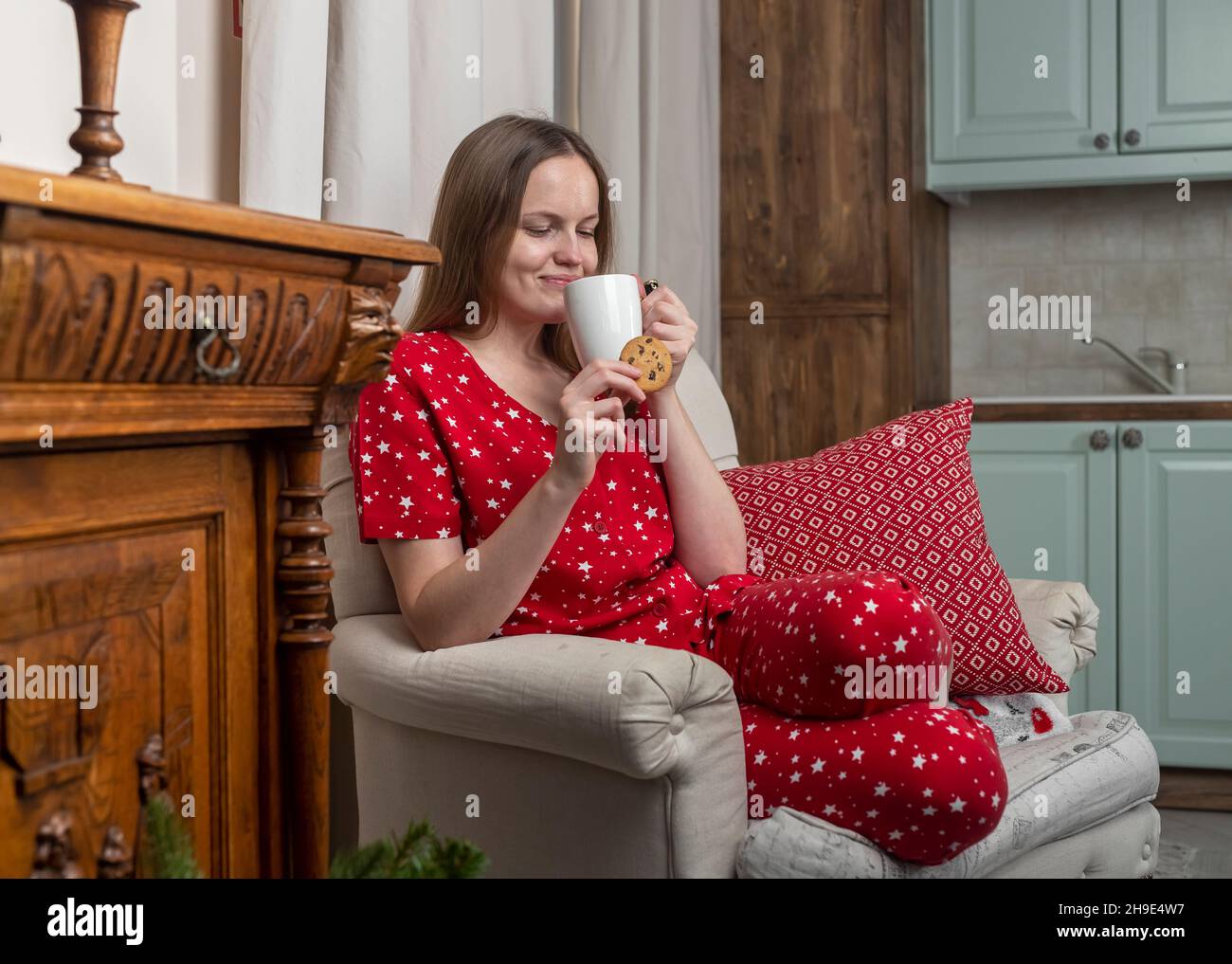 Donna con tazza di tè e biscotti in mani, seduta in sedia a casa accogliente durante le vacanze invernali. Foto Stock