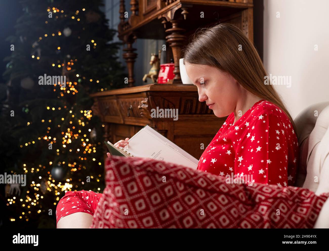 Donna lettura libro, seduta su sedia vicino albero di Natale la vigilia di Natale a casa accogliente. Foto Stock