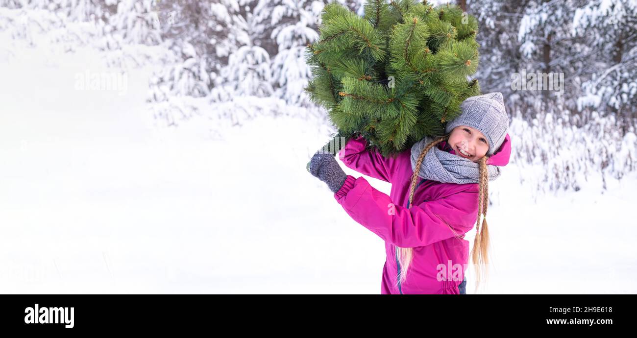 Una ragazza allegra di 10-11 anni in un cappello a maglia e sciarpa che tiene un albero di Natale, sorride, guardando la macchina fotografica contro la neve e gli alberi. Inverno Foto Stock