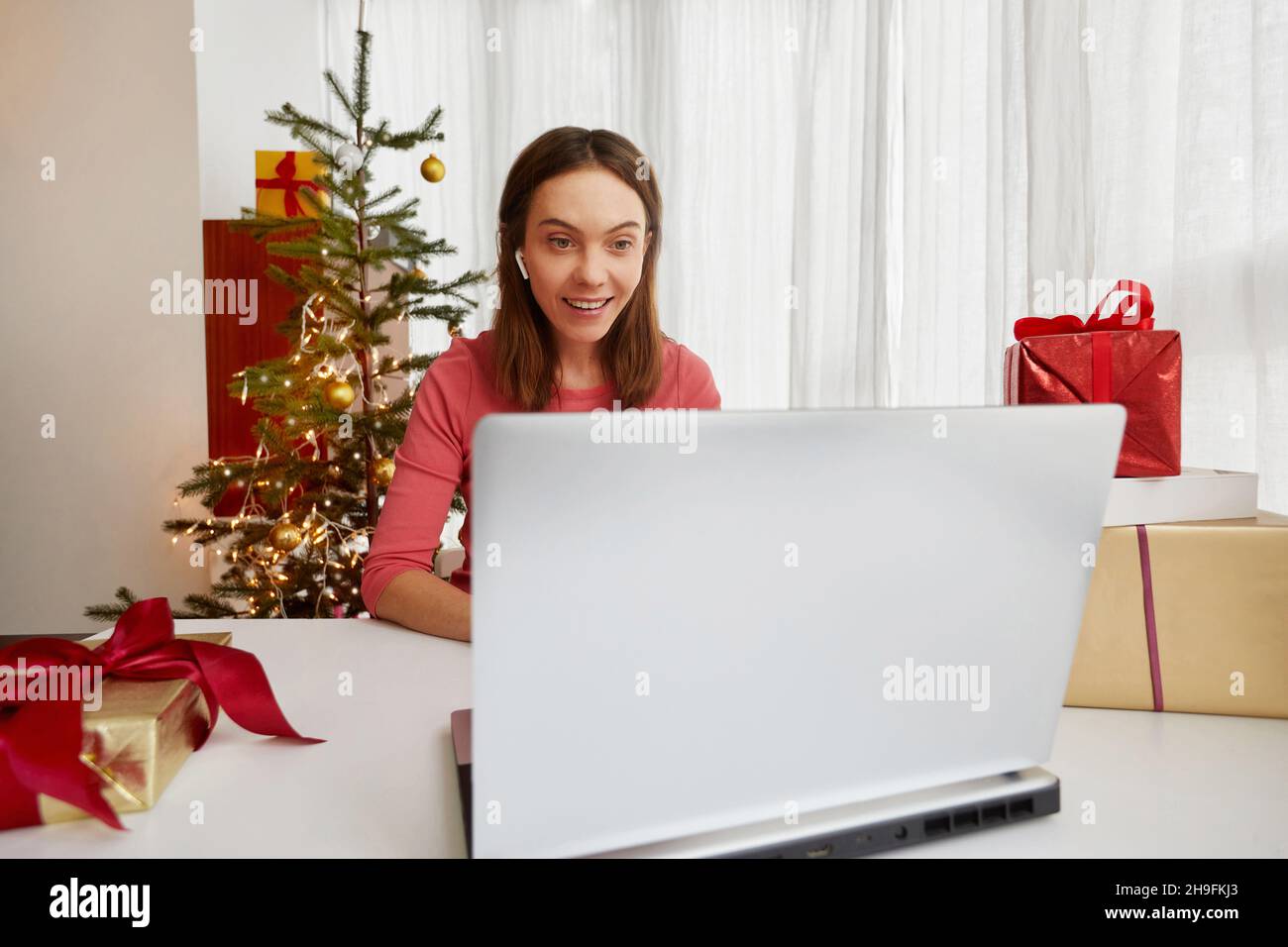 La giovane donna felice siede ad un portatile con le cuffie e l'albero di Natale sullo sfondo tra i regali alla vigilia di Natale Foto Stock