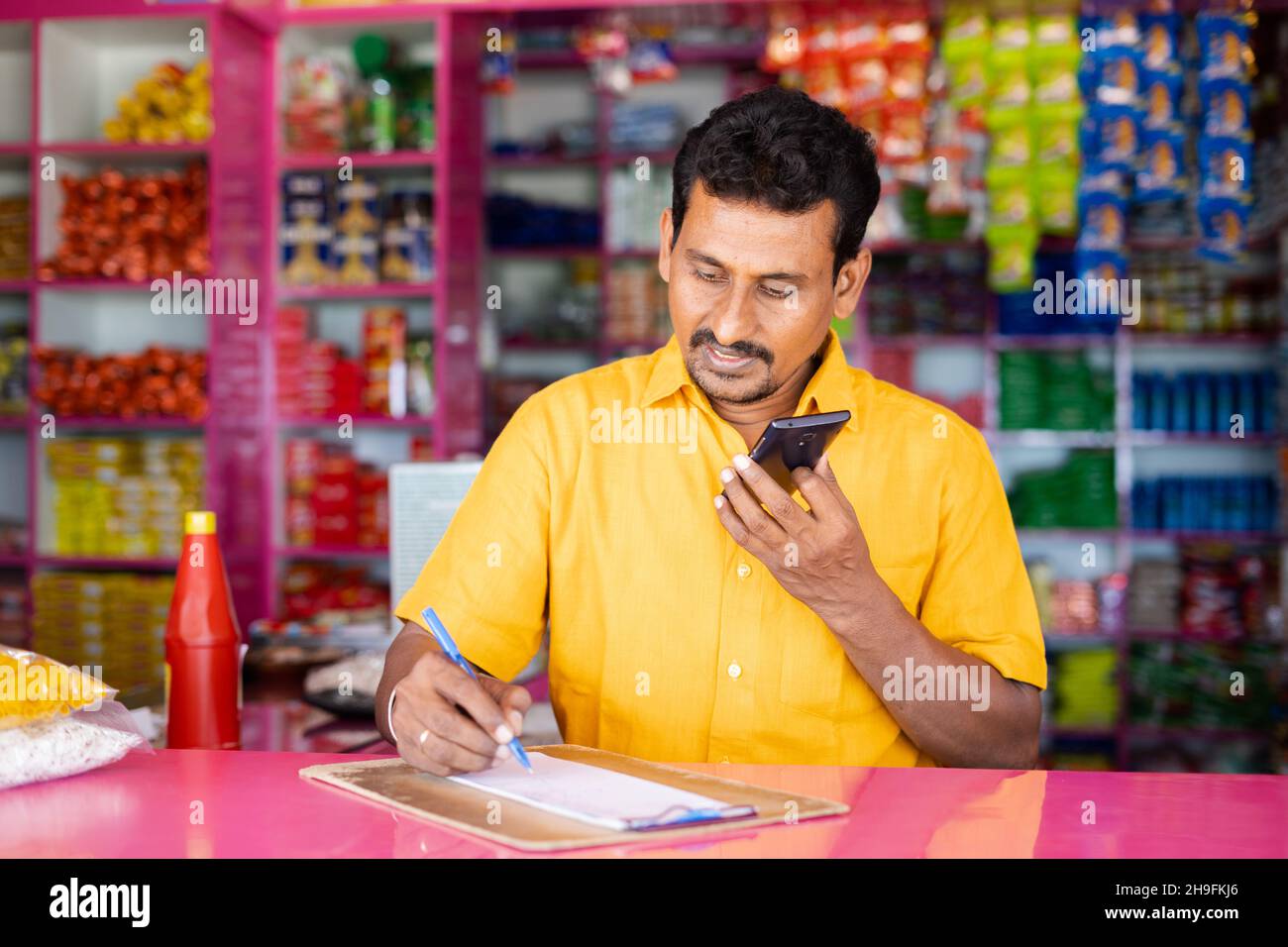 L'uomo d'affari indiano della drogheria che nota gli ordini mentre parla con il cliente sul telefono mobile al negozio di kirana - concetto di shopping a distanza, consegna domestica Foto Stock