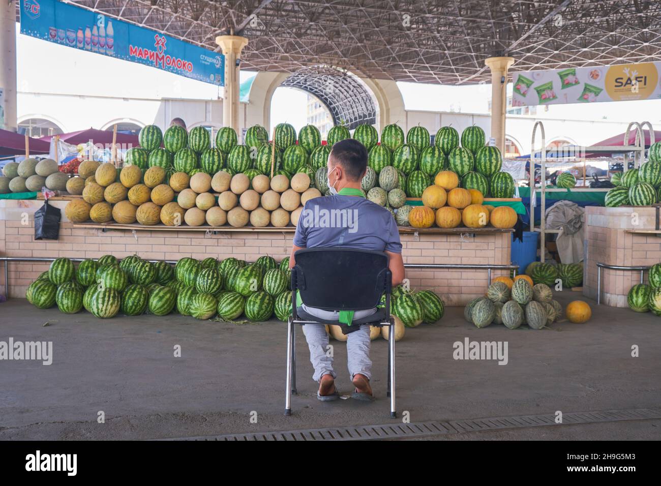 Un venditore in una maschera per Covid che vende cocomero, siluro meloni. Al Bazaar Mirabad, Mirobod Bozori a Tashkent, Uzbekistan. Foto Stock