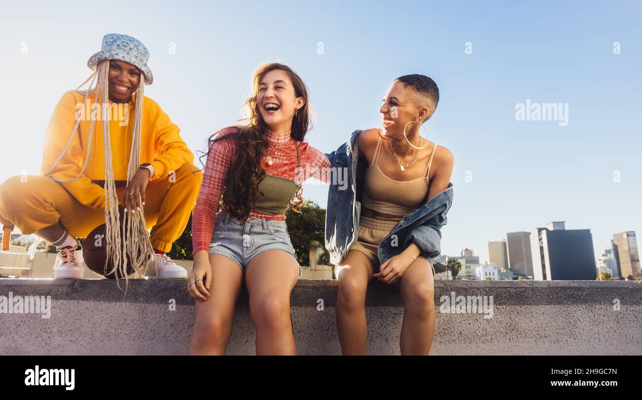 Godendo il tempo della ragazza nella città. Tre amici femminili spensierati ridono allegro mentre si siedono insieme su una parete. I giovani multietnici si divertono Foto Stock