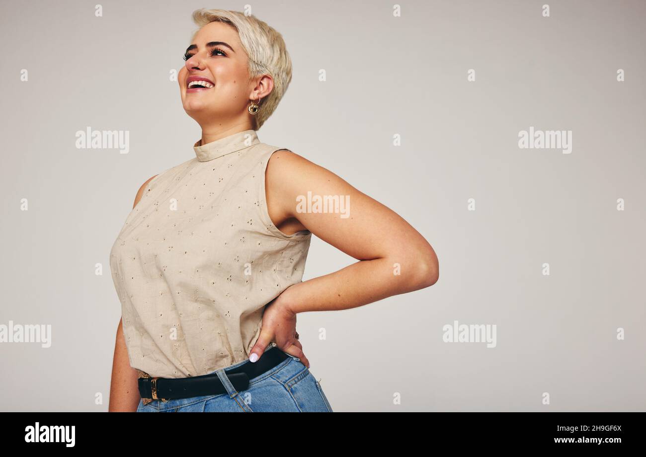 Donna che ride mentre si alza da sola su uno sfondo grigio. Felice donna di metà-adulto che guarda via allegro in uno studio. Donna alla moda che indossa de Foto Stock