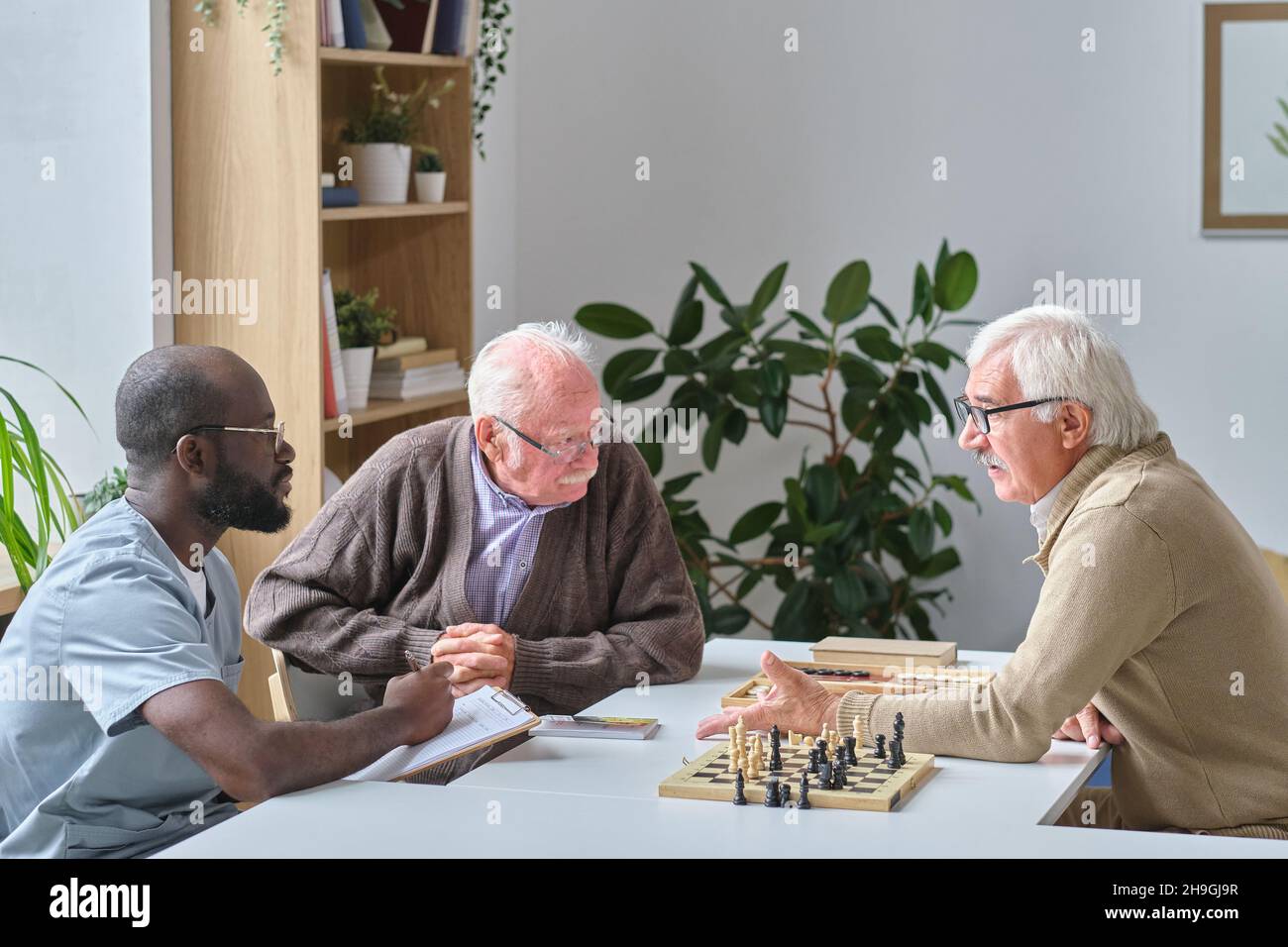 Uomo anziano che parla con il medico africano che si siede insieme a due uomini anziani mentre giocano a scacchi in casa di cura Foto Stock