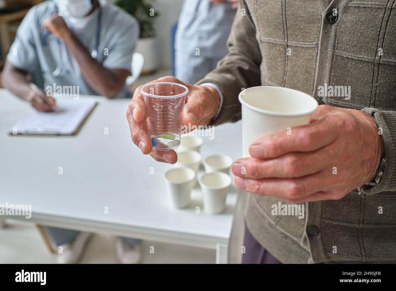 Primo piano di un uomo anziano che tiene un bicchiere d'acqua in una mano e un bicchiere con pillole in altra mano, che prende pillole in ospedale Foto Stock